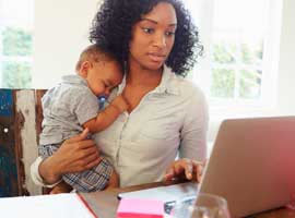 Woman works at kitchen table while holding infant