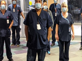 Workers wearing medical gowns, face masks and face shields stand in hallway with fists raised. 