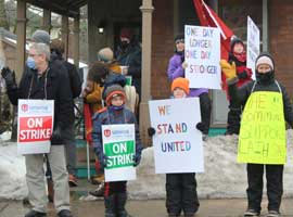 Community members pose on a snow-lined street with signs of support for striking workers.