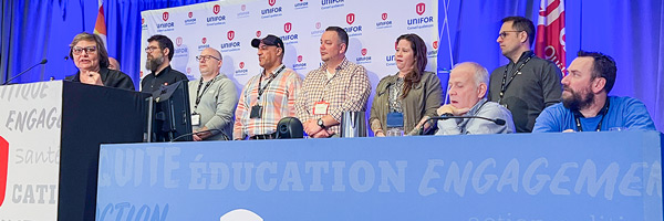 Seven people standing on stage in front of a Unifor Quebec backdrop. One person is speaking from a podium and two are seated.