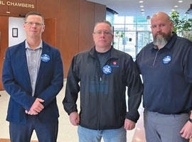 Three men wearing blue buttons standing in front of council chambers. 