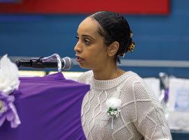 Samia Hashi addresses attendees at podium covered in purple fabric and flowers. 