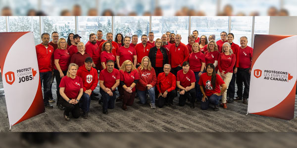 A group of people wearing red shirts stand beside Protect Canadian Jobs signs