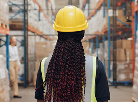 Female warehouse worker wearing hard hat and safety vest looks at shelves of boxes.
